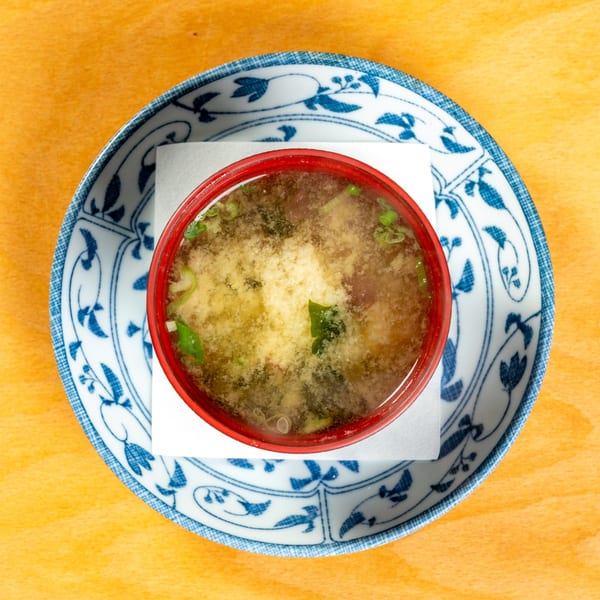 Overhead view of a red bowl of miso soup with green onions and tofu, on a blue and white plate.
